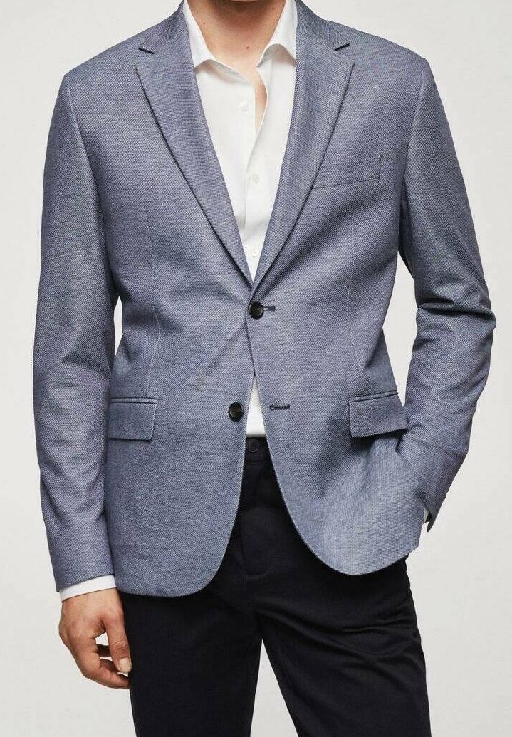 Homme portant un blazer bleu clair texturé sur une chemise blanche et un pantalon noir, une main dans la poche, torse visible, fond neutre.