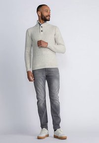 Lichtgrijze gebreide pullover met hoge kraag en drie knopen, gecombineerd met grijze jeans en witte schoenen met een rubberen zool.