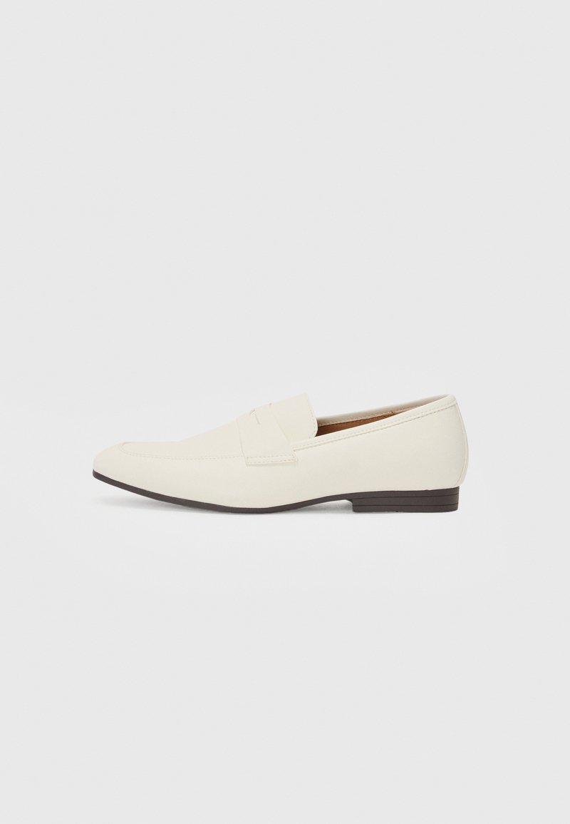 Vaaleanbeige loaferit, joissa on terävä kärki, sileä materiaali, nauha vampin yli ja matala ruskea pohja. Yksinkertainen muotoilu, jossa on minimaalista korostusta.