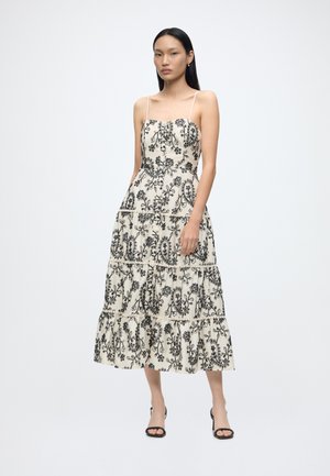 Γυναίκα που φοράει λευκό midi φόρεμα με μαύρα floral μοτίβα paisley και λεπτές τιράντες, στέκεται με το ένα χέρι στην τσέπη και μαύρα σανδάλια με τακούνια.