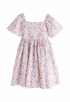 Fată tânără stând cu mâna pe cap, purtând o rochie roz cu flori și mâneci bufante, și sandale bej, pe un fundal simplu.