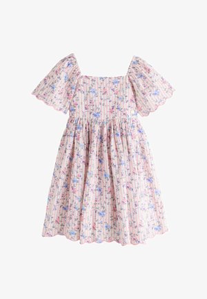 Fiatal lány áll, kezét a fején tartva, rózsaszín virágos ruhát visel, melynek fodros ujjai vannak, és bézs szandált hord, egyszerű háttér előtt.