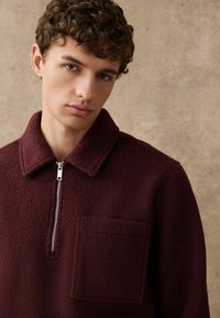 Bordeauxrotes Fleecejacke mit Reißverschluss am Kragen, Kragen-Design, einer Fronttasche und strukturierter Oberfläche. Für eine taillierte Passform entworfen.