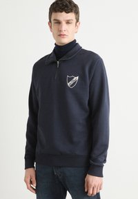 Hanorac bleumarin cu guler cu fermoar pe jumătate, manșete și tiv îmbinate. Prezintă un logo brodat sub formă de scut pe piept. Material din amestec de bumbac.