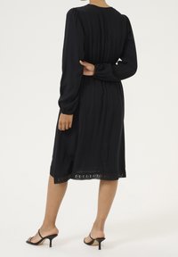 Robe noire à manches longues avec taille cintrée, tissu texturé et bordure en dentelle à l'ourlet. Associée à des sandales noires à talons.