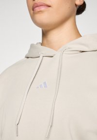 Lichtbeige hoodie gemaakt van zachte stof, met een voorzak, koordjes en een klein paarse logo op de borst.
