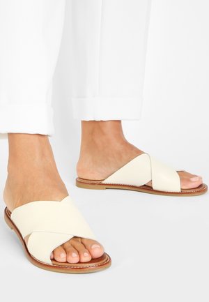 Hvide lædersandaler med krydsede stropper, flad sål og åbne tæer. Glat tekstur med et minimalistisk design.