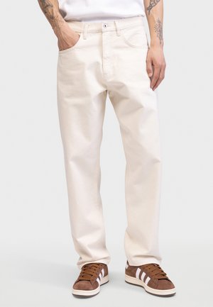 Beige cotton jeans with a straight fit, five-pocket design, and visible stitching, paired with brown suede sneakers featuring white stripes.