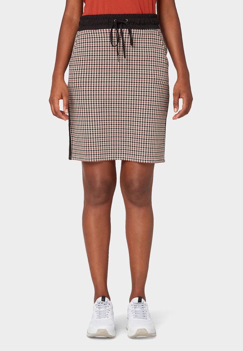 Femme portant une jupe pied-de-poule longueur genoux avec taille à cordon noir, haut orange et baskets blanches sur fond uni.