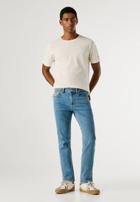 Beżowa bawełniana koszulka z krótkim rękawem, zestawiona z jasnoniebieskimi dżinsami i beżowymi trampkami z czarnymi i białymi akcentami.