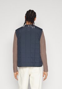 Marinblå quiltad väst med ärmlös design och texturerad tyg. Har tryckknappslås vid halsen och detaljerad sömnad över hela vesten.