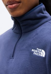 Marineblauwe fleece pullover met een hoge kraag, halve ritssluiting en een wit geborduurd logo op de borst. Zachte textuur en een gladde afwerking.