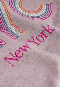 Gros plan sur un tissu violet clair avec des lettres brodées colorées formant une partie du mot "New York" en fil rose.