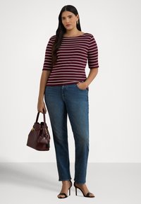 Sieviete valkā bordo un baltas svītras augšdaļu, zilas džinsas, melnas kurpes ar papēžiem un rokās tur saskaņotu bordo rokassomiņu ar pievienotu mazu piederumu somiņu.