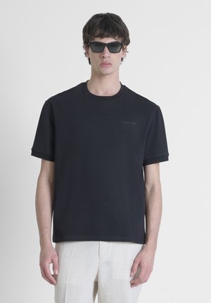 Camiseta negra de manga corta en algodón con cuello redondo y un sutil detalle de logotipo grabado en el pecho. Llevada con pantalones de color claro.