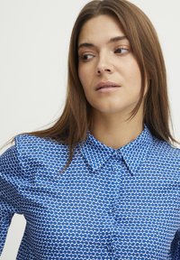 Camicia azzurra con bottone, caratterizzata da un motivo a onde bianche. Presenta un colletto classico e un tessuto leggermente texturizzato.