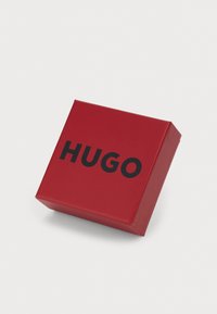 Rød firkantet emballasje med blank finish, som har en fremtredende svart "HUGO"-logo på toppen.