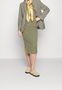Midikleid in Olivgrün mit einem seitlichen Schlitz, kombiniert mit einem hellgrauen Blazer und beigen Slippern. Dazu ein gelber Seidenschal.