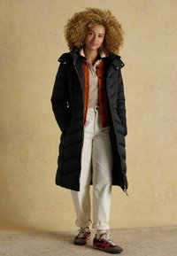 Melna quiltota garā mētelis, oranžs pogājamais džemperis virs bēša augšdaļas, gaišu piedurkņu bikses un brūni sporta apavi ar sarkaniem akcentiem.