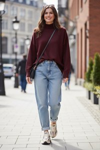 Mujer joven caminando por la acera de la ciudad con un suéter burdeos, vaqueros azul claro, zapatillas deportivas y gafas de sol en la cabeza.