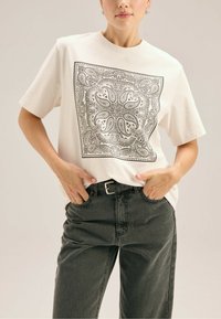 Biała bawełniana koszulka z czarnym wzorem paisley w kszta�łcie kwadratu; noszona z ciemnoszarymi, wysokim stanem jeansami i czarnym paskiem.