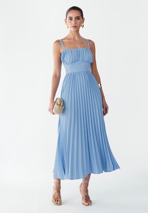 Žena v svetlomodrých plisovaných midi šatách bez rukávov, drží béžové tkané psaníčko, nosí zlaté sandále na remienkoch a kruhové náušnice.