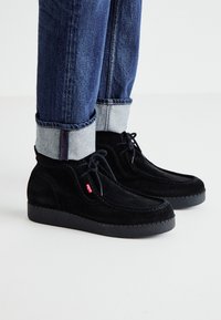 Chaussures en daim noir avec une finition texturée, bout rond et semelle en caoutchouc noir contrastante. Dotées de lacets et d'une petite étiquette de logo rouge.