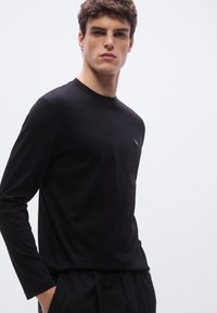 Musta pitkähihainen paita, jossa on pyöreä pääntie ja sileä kangas. Vasemmassa ylärinnassa pieni logo.