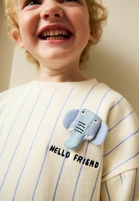 Krémový svetr s modrými svislými pruhy, s aplikací světle modrého slona a černým vyšitým textem "HELLO FRIEND."