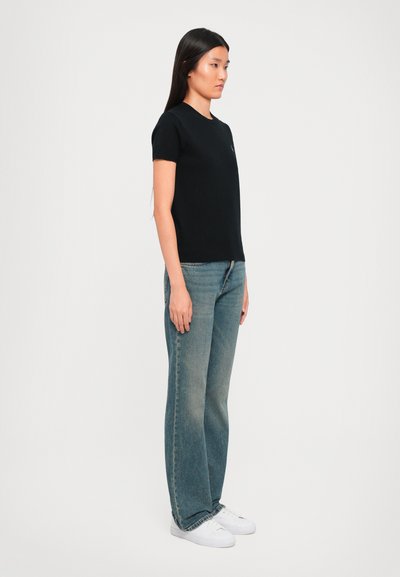 Woman standing in profile wearing a black fitted t-shirt, blue jeans, and white sneakers against a plain white background.