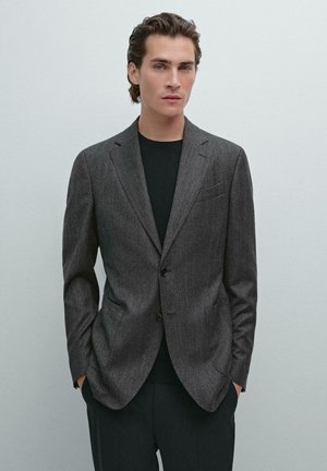 Gray tailored blazer made of textured fabric, single-breasted with two buttons, featuring front pockets and a notch lapel, worn over a black shirt.