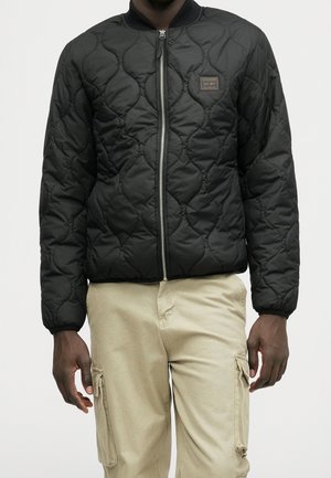 Homme portant une veste matelassée noire à fermeture éclair et un pantalon cargo beige, visible du cou aux hanches sur un fond uni.