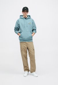 Hanorac albastre deschis cu buzunar frontal și logo, purtat cu pantaloni cargo khaki, prevăzuți cu buzunare laterale, completat cu adidași albi și o șapcă neagră.