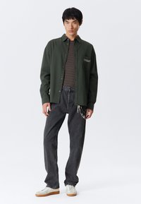 Young man standing wearing dark green button shirt over brown striped tee, loose black jeans with hanging chains, and white sneakers.
