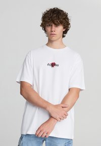 Biała bawełniana koszulka z krótkim rękawem i swobodnym krojem. Posiada czarny napis oraz grafikę czerwonej róży na klatce piersiowej.