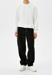 Vit långärmad sweatshirt med rund halsringning och sömdetaljer, tillsammans med svarta cargobyxor med vit sömnad och fickor.