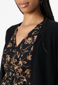 Cardigan nero su un vestito floreale scuro con scollatura a V, dotato di motivi floreali dorati e marroni e una vita aderente con dettaglio dei bottoni.