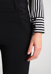 Mano destra appoggiata di lato, indossa pantaloni neri con tasca e una camicia a righe verticali bianco e nera con i polsini.