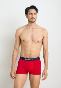 Boxers rouges en tissu extensible, offrant un ajustement serré et une taille contrastante bleu marine avec le logo "LACOSTE" imprimé.