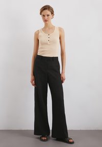 Beige geribbelde tanktop met drie bruine knopen, gecombineerd met hoge zwarte wijde broek en zwarte slipper sandalen.