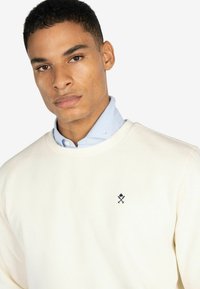 Suéter de crema claro con cuello redondo, con detalle de logo bordado en negro y llevado sobre una camisa de color azul con cuello. Textura de tela suave.