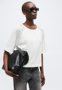 Γυναίκα με πλεξούδες που φοράει μαύρα γυαλιά ηλίου, λευκό μπλουζάκι με κοντά μανίκια, γκρι τζιν και κρατά μια μεγάλη μαύρη διπλωμένη δερμάτινη τσάντα.