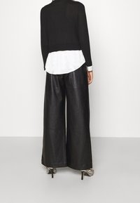Persona che indossa pantaloni in pelle neri a gamba larga, maglione nero sopra una camicia bianca e tacchi a zebra, in piedi di fronte a un muro bianco.