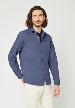 Chemise à rayures bleues marine avec une texture douce, dotée d'un col, de manches longues et d'un petit logo sur la poitrine. Assortie à un pantalon crème.