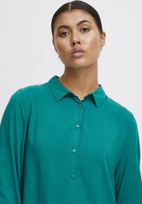 Teal blouse with a collared neckline, long sleeves, and button-down front. Made of smooth fabric with a subtle sheen.