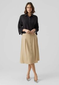 Schwarze Hemdbluse mit weiten Ärmeln, kombiniert mit einem beigen, plissierten Midirock mit Knöpfen an der Vorderseite. Getragen mit metallischen High Heels.