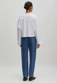 Weißes Baumwolloberteil mit langen Ärmeln und Rüschendetails, kombiniert mit blauen, hoch geschnittenen Jeans. Einfaches Design mit lockerem Schnitt und Rückansicht.
