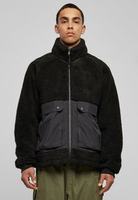 Giacca in pile nero ad alto pelo con zip frontale, caratterizzata da una parte inferiore in nylon nero liscio, due tasche con bottone a pressione e colletto alto.