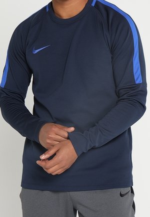 Persoon draagt een marineblauw Nike sportshirt met lange mouwen, blauwe strepen op de schouders en grijze broek met een zwart Nike-logo.