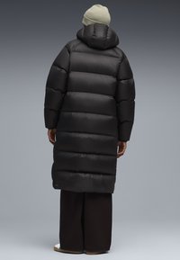 Zwarte pufferjas met een capuchon, heeft geblokte secties, lange lengte en een gladde textuur. Gecombineerd met donkere broeken en een lichte beanie.